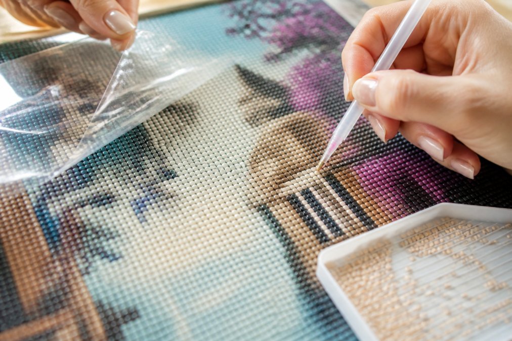 Close-up of hands carefully placing resin diamonds onto a canvas with poured glue, showing the sticky surface with a clear protective cover partially peeled back, natural studio lighting