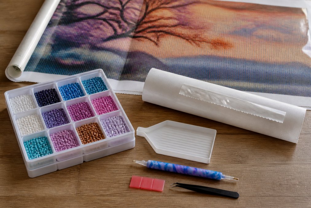 Flat lay of a premium diamond painting kit displaying specialized drill pen with ergonomic grip, diamond tray with multiple compartments, roll of release paper, and canvas showing original artwork pattern with clear protective cover