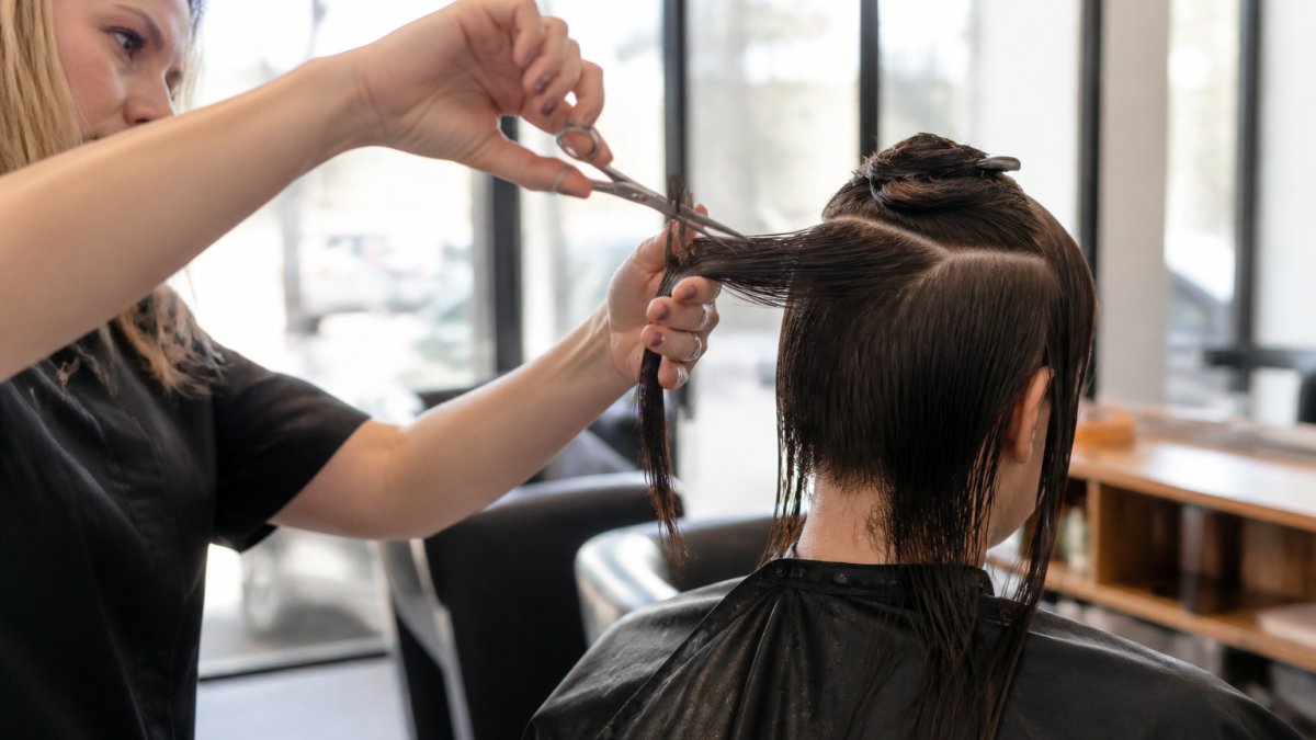 A stylist's hands holding sharp shears mid-cut on a client's damp hair in a bright, upscale salon, with clean sectioned strands visible under soft window light