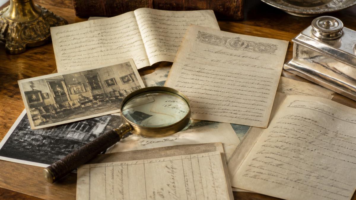 Close-up of antique documents, receipts, and handwritten letters spread on a wooden desk with a magnifying glass, showing historical ownership records and provenance materials