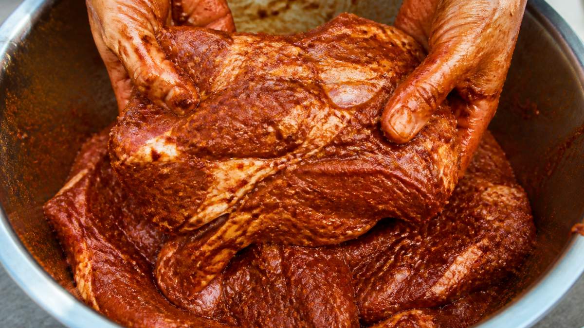 Hands coating raw chuck roast with deep red chile marinade paste, close-up showing the spice coating on raw beef in a large bowl