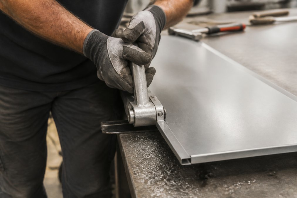 Professional roofer using a hand seamer tool to create a precise 90-degree bend on a metal panel edge, with the tool positioned perpendicular to the panel and metal shavings visible from the bending process