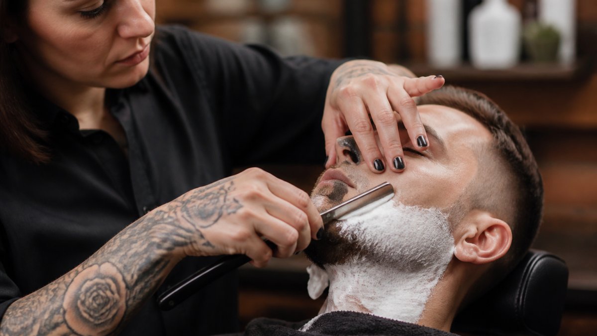 Professional barber demonstrating proper straight razor grip and blade angle positioning against client's face with steady hand positioning and correct 30-degree angle alignment