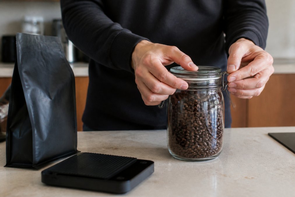 A person's hands sealing an airtight glass jar filled with whole roasted coffee beans on a kitchen counter, with a digital scale and a bag of beans nearby, warm natural morning light