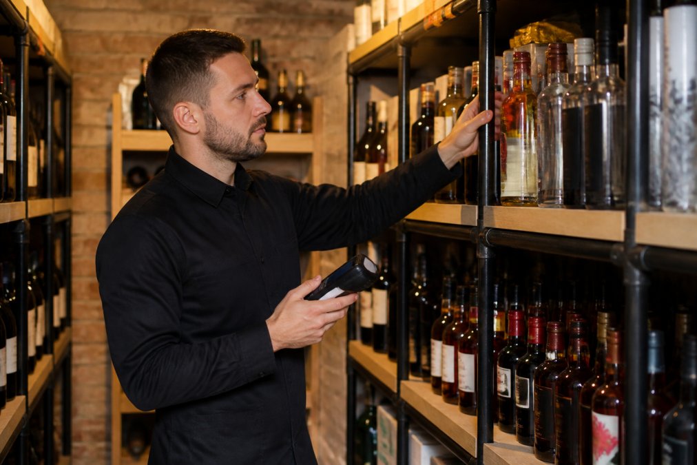 A bar manager using a digital stocktaking device to count bottles on shelves in a well-organised cellar, with warm ambient lighting