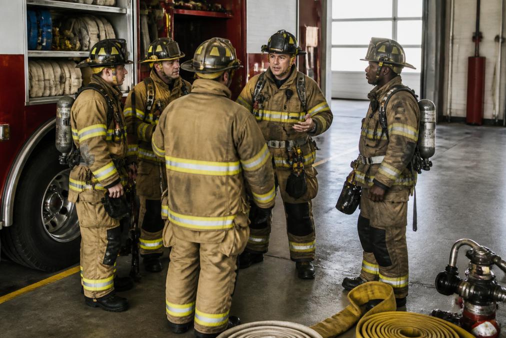 A fire crew in full turnout gear and helmets gathered around a truck at a station bay, talking through a plan before a drill, with hose and tools visible nearby