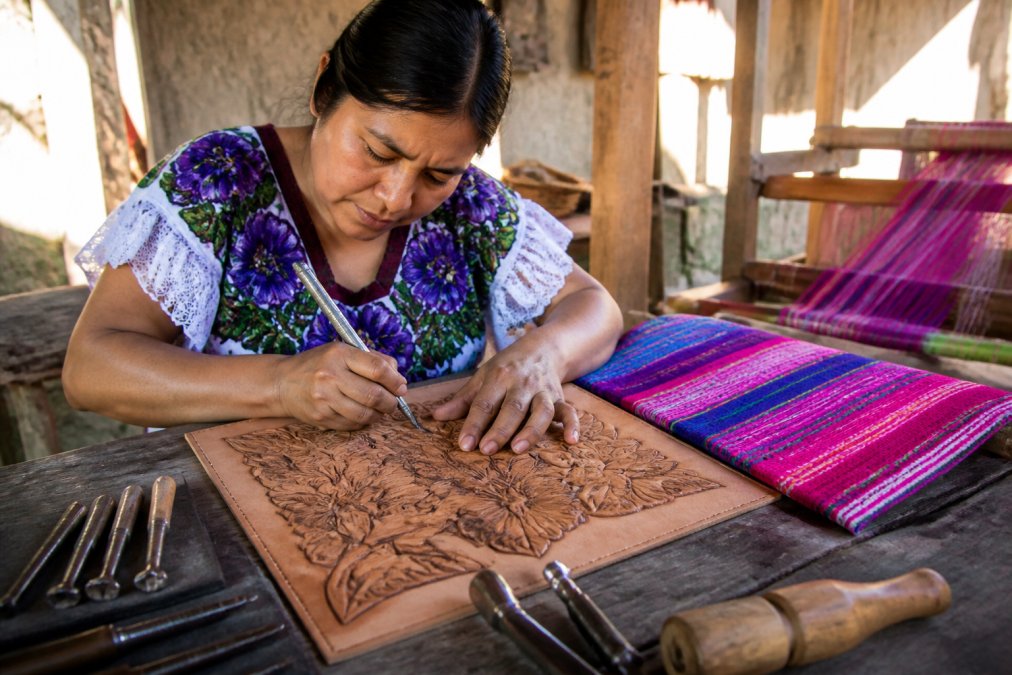 Artisan carving floral leather designs for fair trade accessories in a bright, traditional workshop.