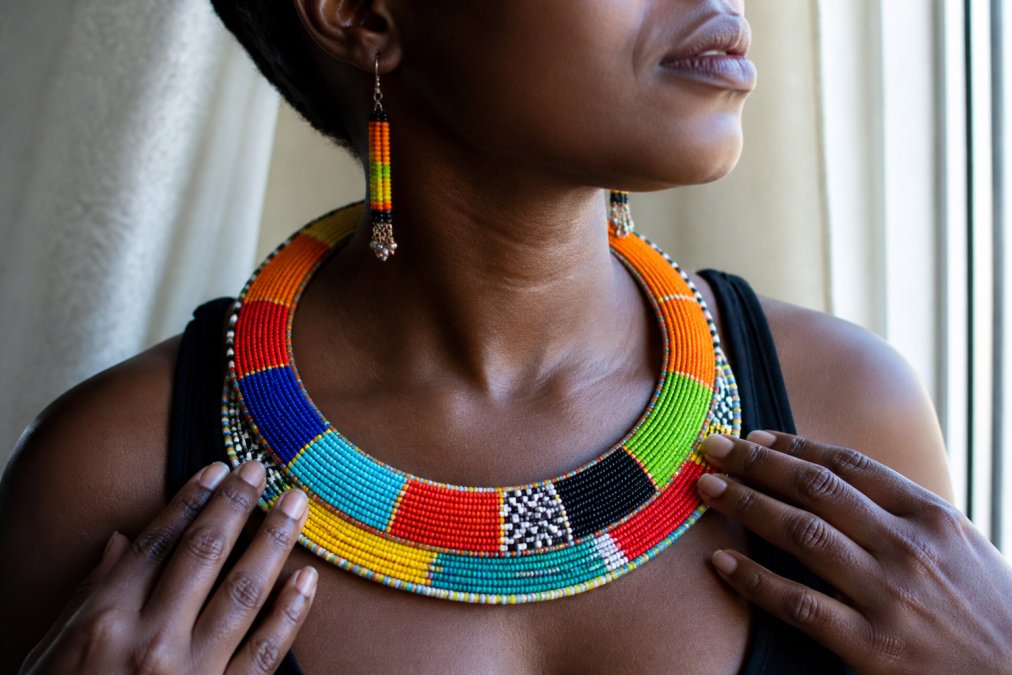 Close-up of a woman's neck and hands wearing a vibrant, colorful chunky beaded African tribal necklace and matching earrings, soft natural window light catching the texture of each bead
