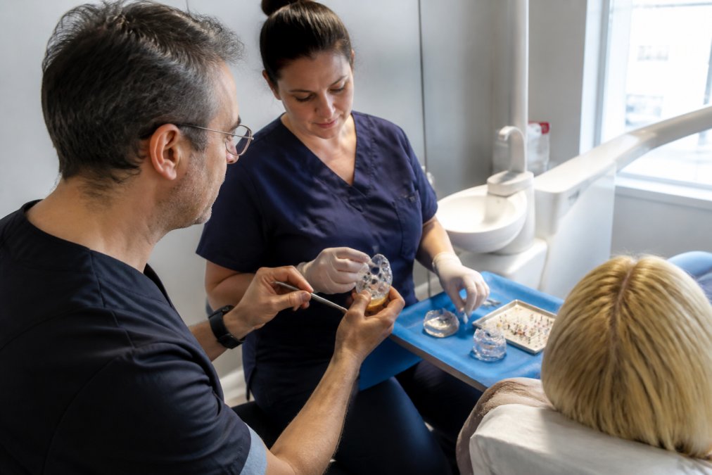 A dentist in a bright modern surgery reviewing a 3D digital scan of a patient's jaw on a screen, with a dental nurse beside them preparing guided surgery equipment
