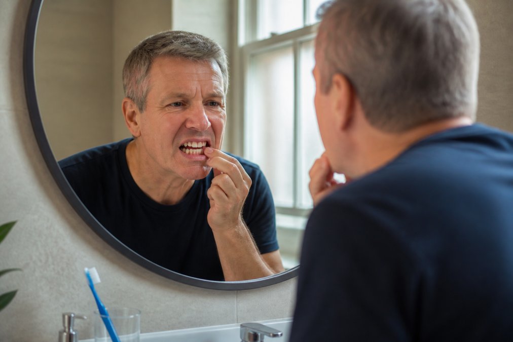 A person in their fifties examining their gum line in a bathroom mirror with a soft toothbrush on the counter, warm natural light, calm and health-conscious mood