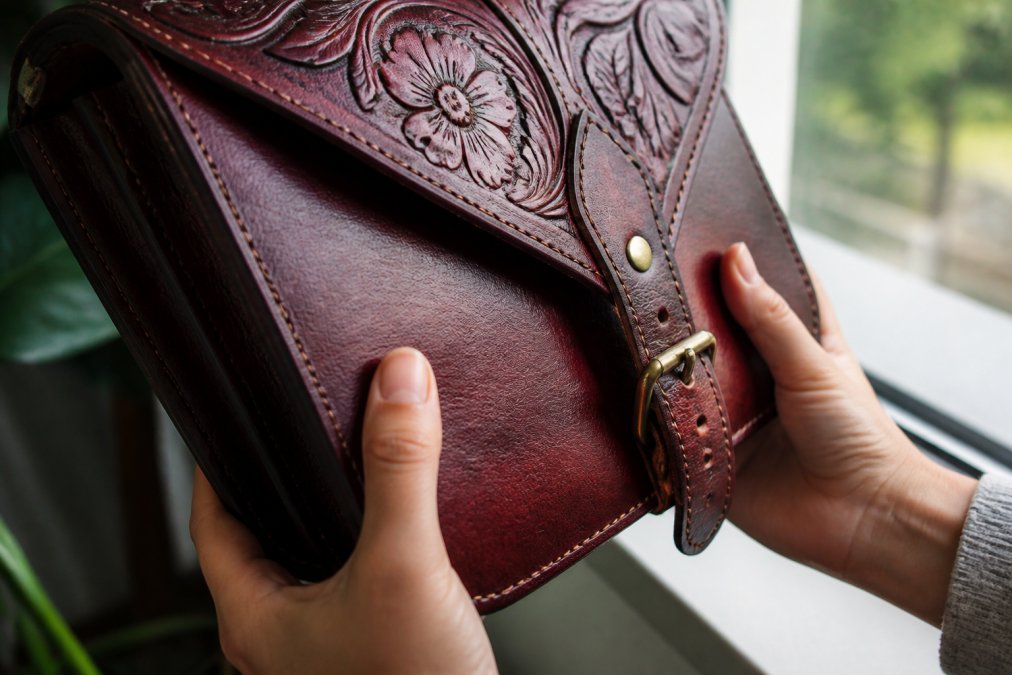 Close-up of hands examining the natural grain texture of a burgundy leather satchel in bright window light