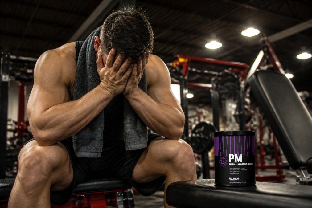 Fatigued bodybuilder sitting on gym bench with head in hands, shoulders slumped, towel draped over neck, harsh gym lighting, clearly exhausted and mentally drained