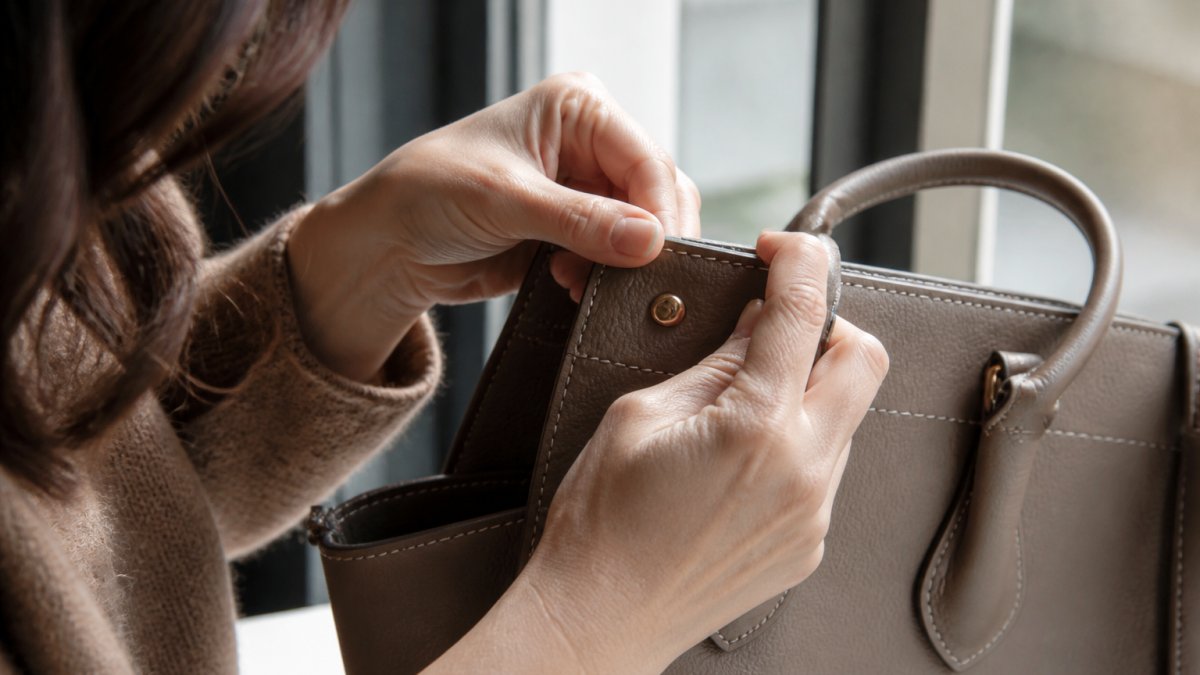 Close-up of a woman's hands inspecting the stitching and edge paint on a structured leather handbag in natural window light