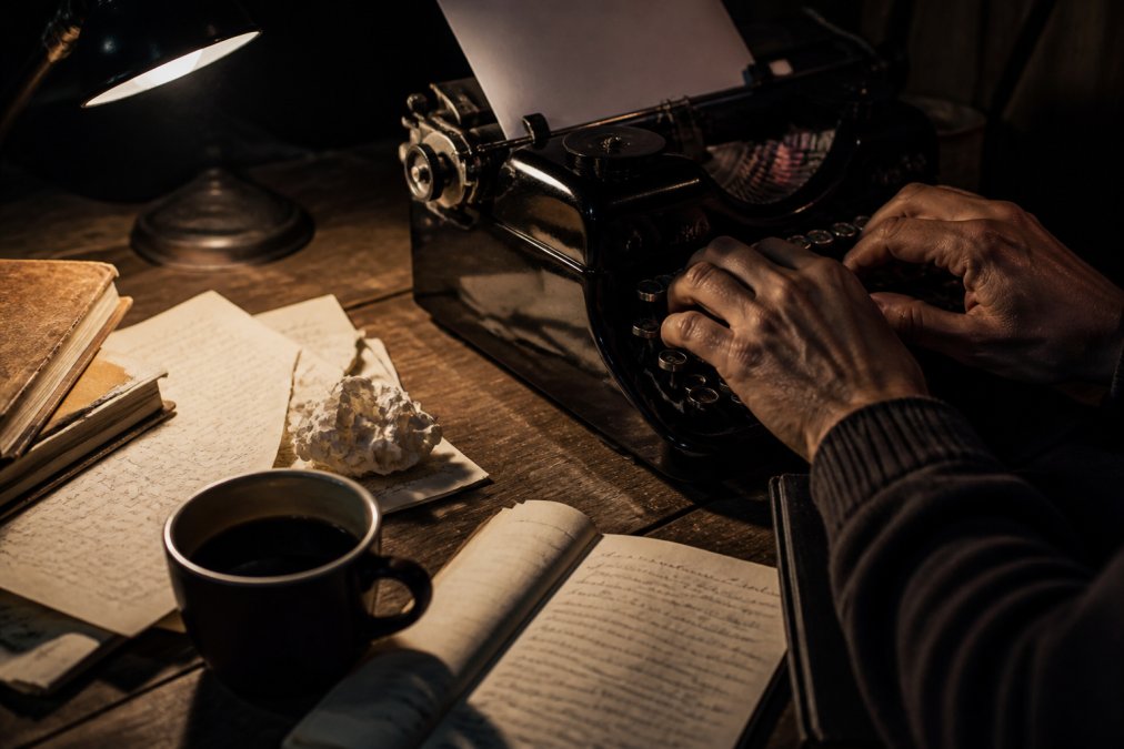 A close-up of a writer's hands typing on a vintage typewriter, with scattered handwritten notes and a cup of coffee on a wooden desk, moody lamplight casting long shadows