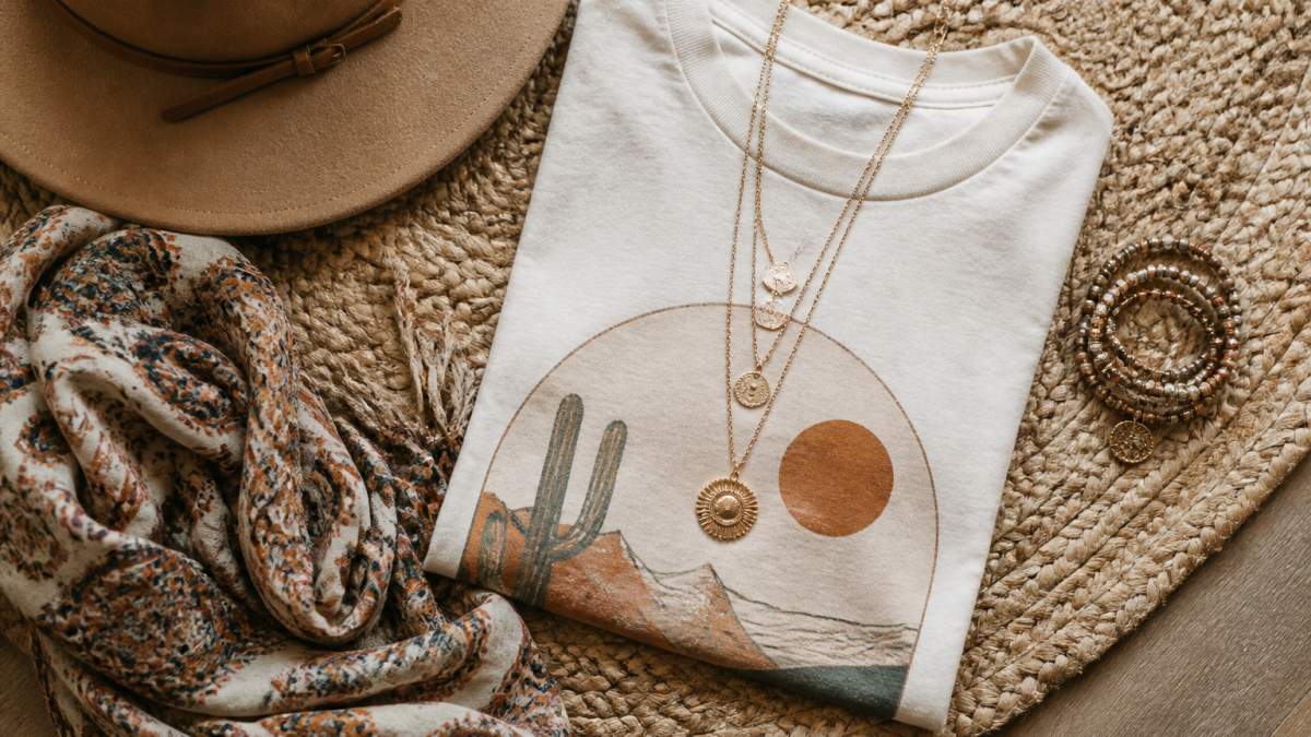 Flat lay of boho accessories on a woven rug, layered gold necklaces, a wide-brim hat, a patterned scarf, and stacked bracelets arranged next to a folded graphic tee in soft natural light