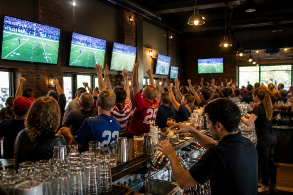 A packed sports bar with multiple large high-definition televisions mounted on walls, diverse fans cheering while watching a live game, warm ambient lighting, bartenders pouring drinks in the foreground