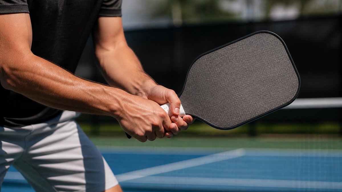 Professional player's hands gripping a high-performance pickleball paddle mid-volley on an outdoor court, showing paddle face texture and grip detail in natural sunlight