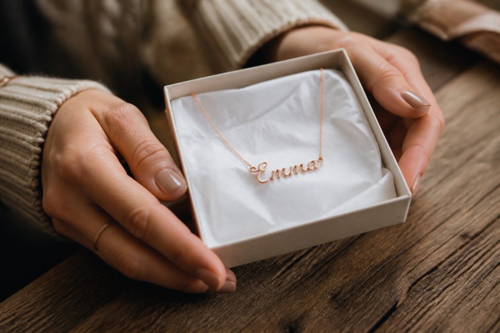Close-up of a woman's hands holding a personalized name necklace nestled in a small gift box lined with white tissue paper, warm soft lighting on a weathered wooden table