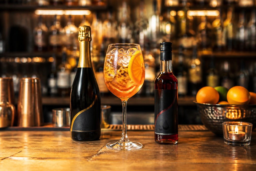 An elegantly crafted alcohol-free Spritz cocktail with Pierre Chavin Sparkling Chardonnay and Anon Bittersweet, garnished with fresh citrus and ice, in a wine glass on a wooden bar counter with warm ambient lighting