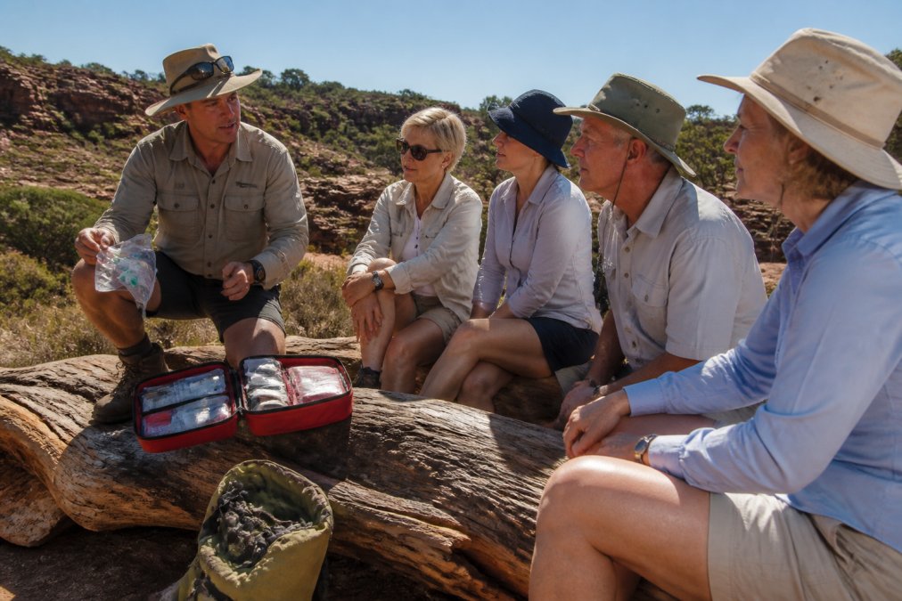 A tour guide in outdoor gear kneeling beside an open first aid kit and demonstrating its contents to a small group of attentive travellers at a remote bush campsite, rocky terrain and scrub visible in the background under clear midday light