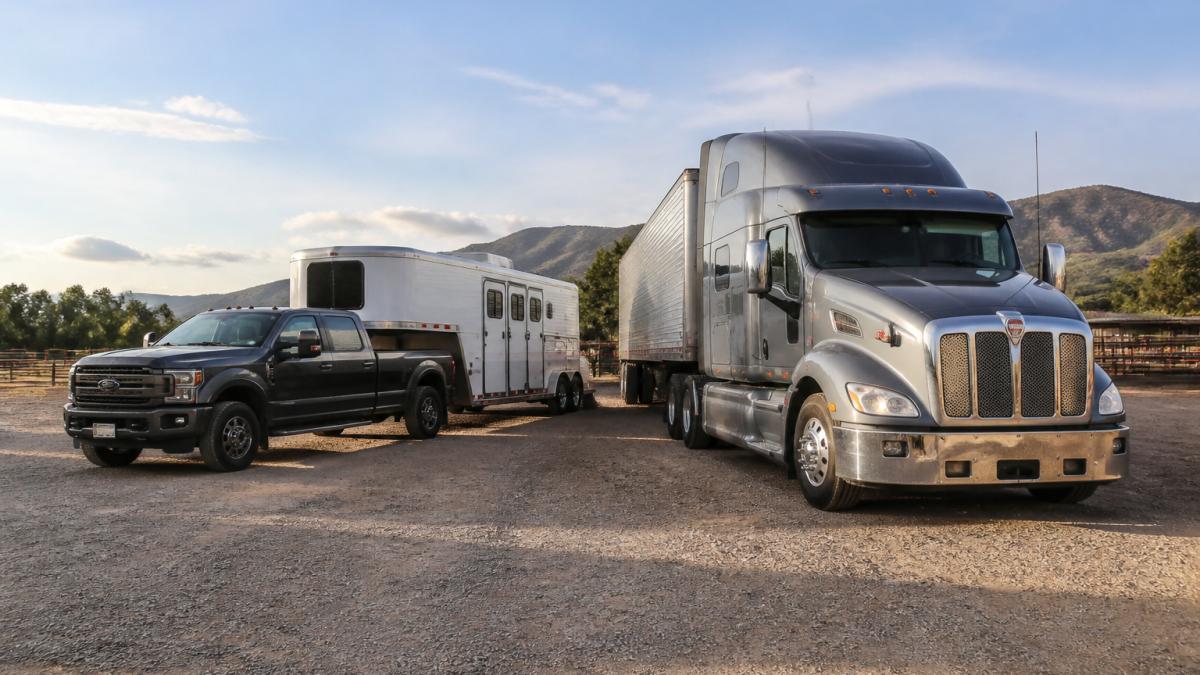 A heavy-duty pickup truck with a gooseneck horse trailer parked beside a large Class 8 semi-truck at a rural staging area in warm afternoon light, showing the size difference between the two vehicles
