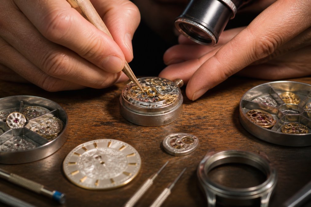 Watchmaker's hands performing precision finishing work on a custom watch movement under magnification, with polishing tools, jeweled bearings, and ornate dial components visible on a workbench