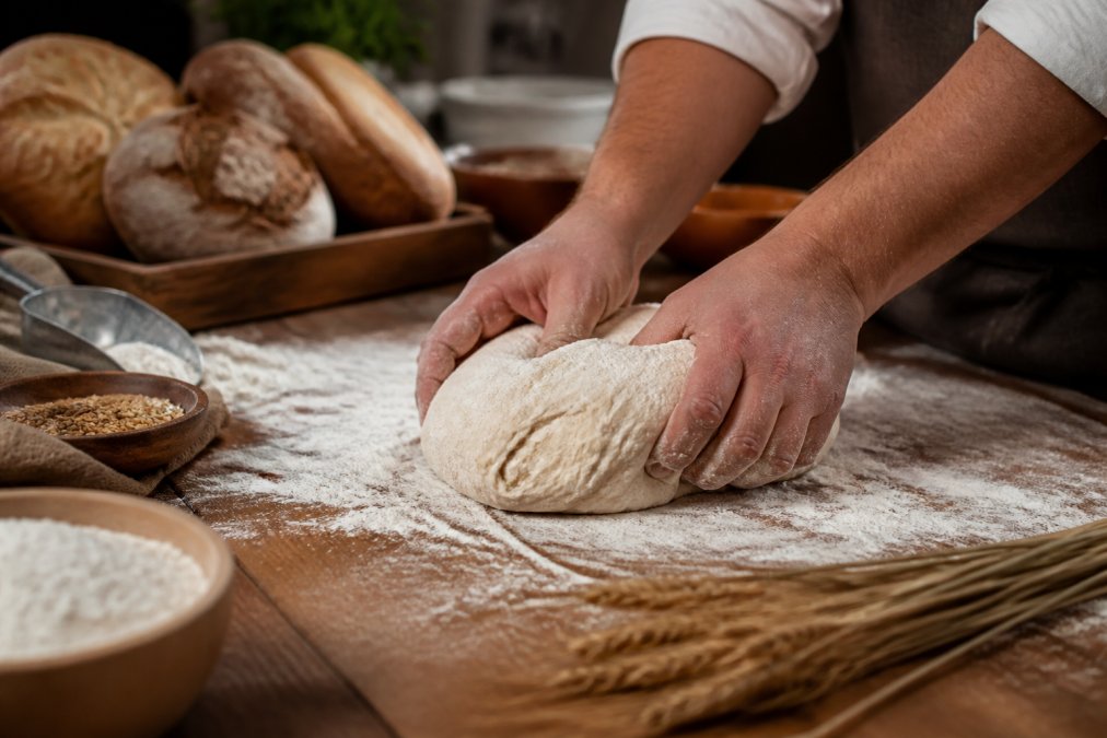 Close-up of a baker's hands kneading dough on a flour-dusted wooden table, with artisan bread loaves and raw ingredients softly blurred in the background