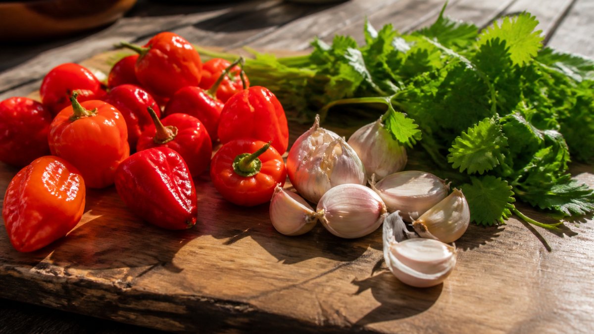 Close-up of vibrant red and orange habanero peppers, fresh garlic cloves, and green cilantro arranged on a rustic wooden cutting board with morning sunlight casting shadows across the ingredients