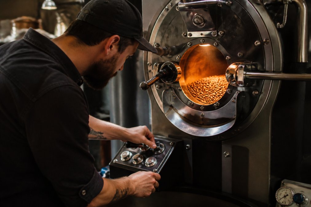 Skilled roaster monitoring coffee beans in a small-batch roaster, warm amber light illuminating the roasting chamber with beans mid-roast, hands adjusting controls with focus and precision