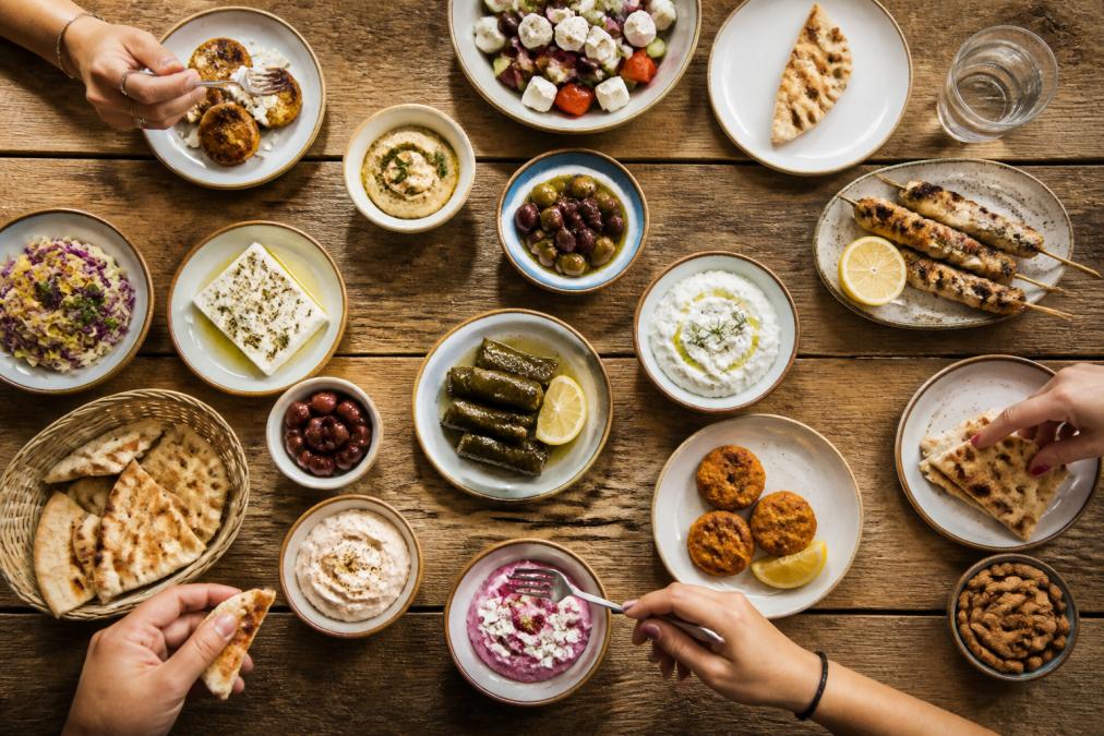 Overhead shot of a rustic wooden table filled with small plates of Greek meze including olives, feta, dolmades, tzatziki, and warm pita bread, with hands reaching in to share