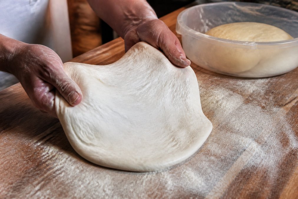 Hands stretching a fermented dough ball on a floured counter to create high quality pizza with a rising bowl nearby.
