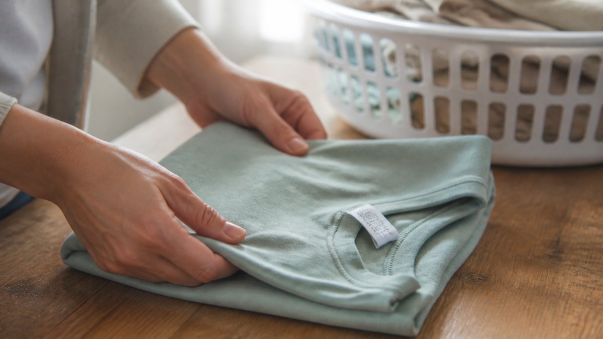 Close-up of hands folding a soft cotton t-shirt on a wooden table, with a care label visible and a laundry basket in the background