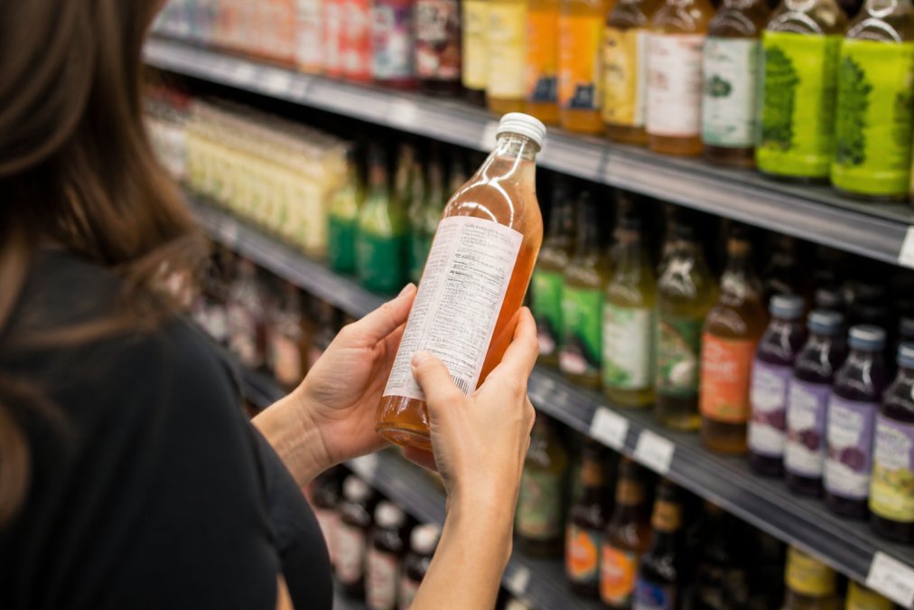 A person's hands holding a glass bottle of kombucha in a grocery store aisle, turning the label to read the ingredients list, with other bottled beverages blurred in the background