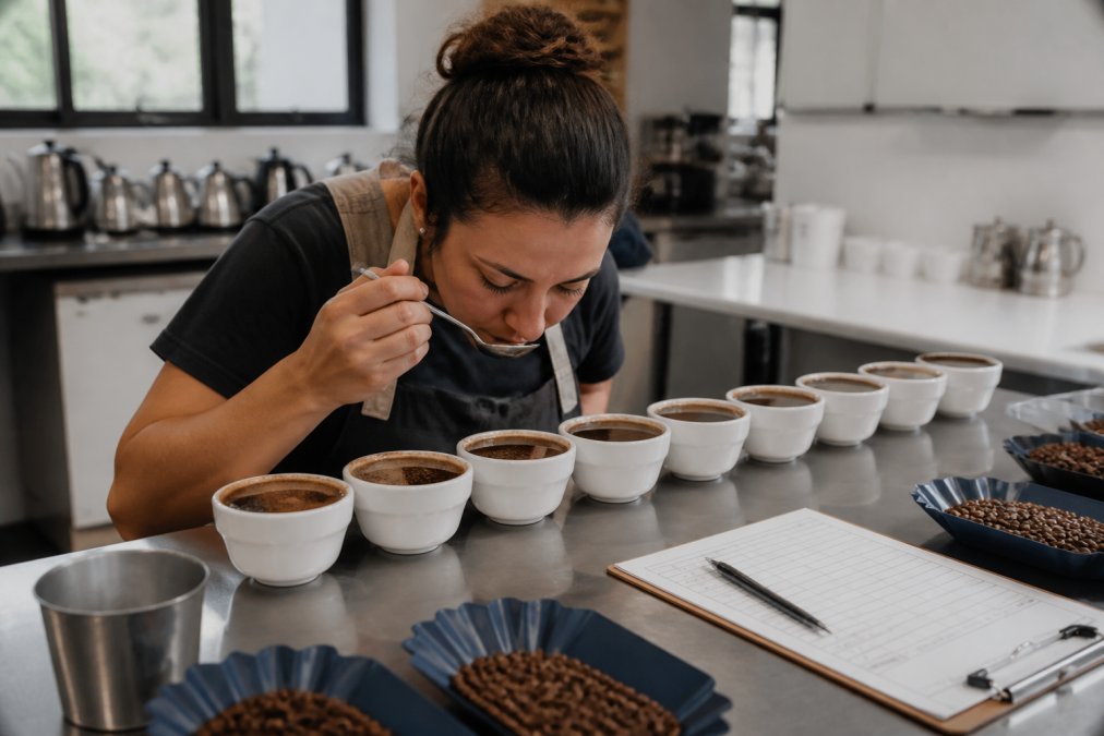 A coffee grader at a cupping table, spoon in hand, leaning over a row of white cups filled with dark coffee, with a scoring sheet and pen nearby in a bright professional cupping lab