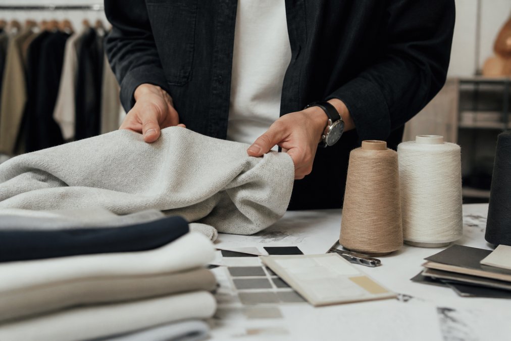 Close-up of hands examining fabric texture, feeling the weight and drape of a premium cotton garment on neutral background with natural lighting
