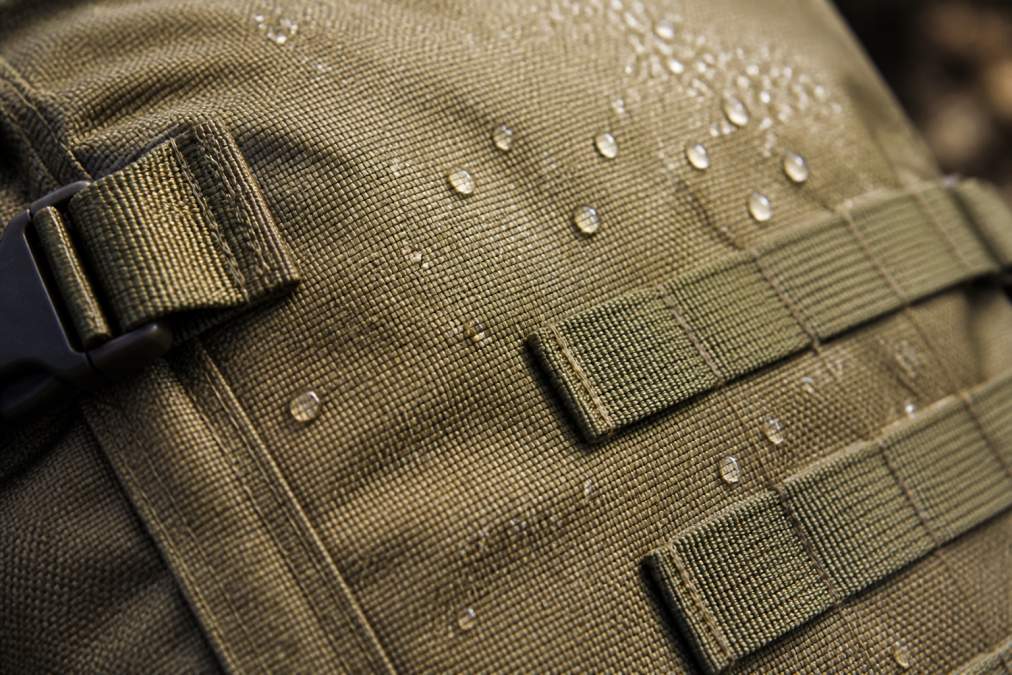 Close-up of a rugged tactical backpack fabric showing tight ripstop weave, reinforced bar-tack stitching, and a water-repellent finish with water beading on the surface