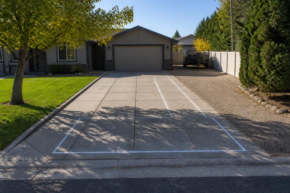 Wide residential driveway with clear space marked by chalk or tape showing the delivery zone, access path visible from street to driveway without overhead wires, tree branches, or power lines obstructing the space, morning sunlight casting clear shadows