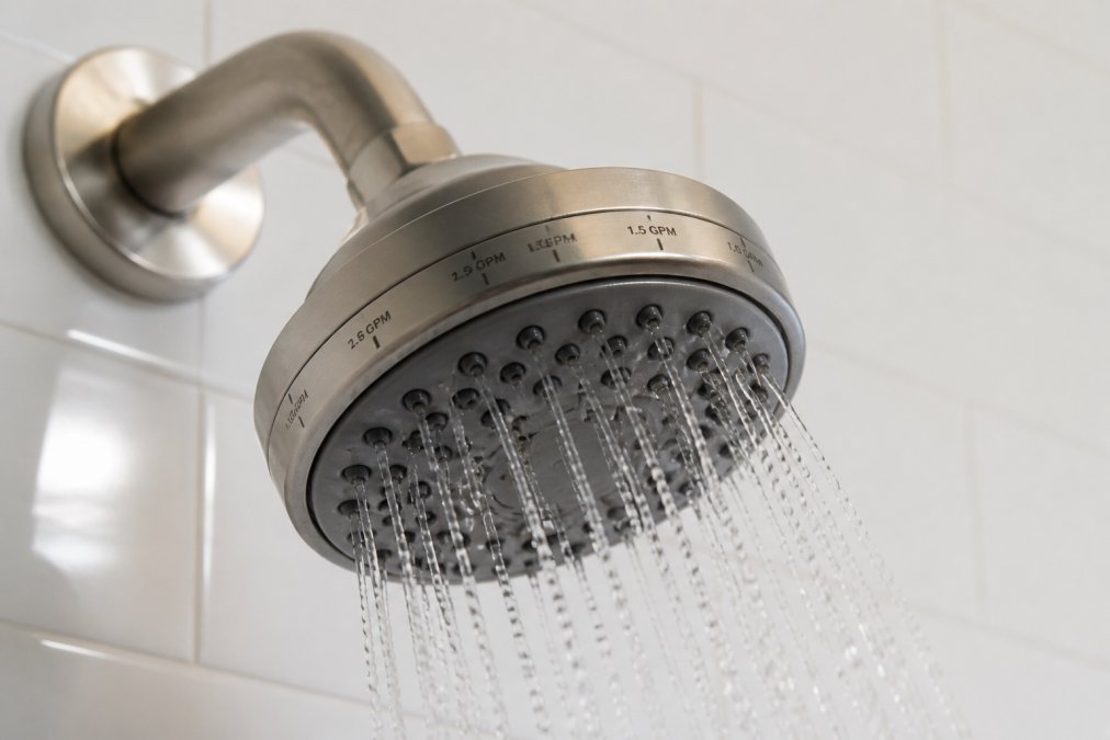 Close-up of a showerhead with flow rate markings visible on the collar, water droplets falling, mounted on a white tile wall with natural bathroom lighting