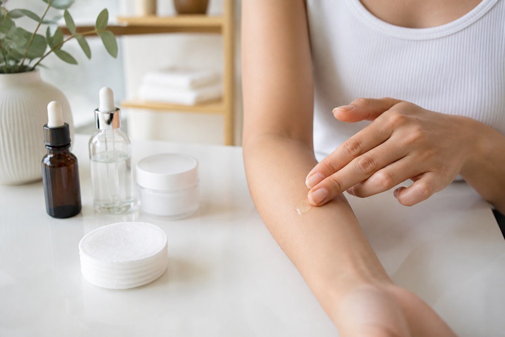 Close-up of hands applying a small amount of serum to the inner arm for patch testing, with skincare products arranged nearby on a clean white surface