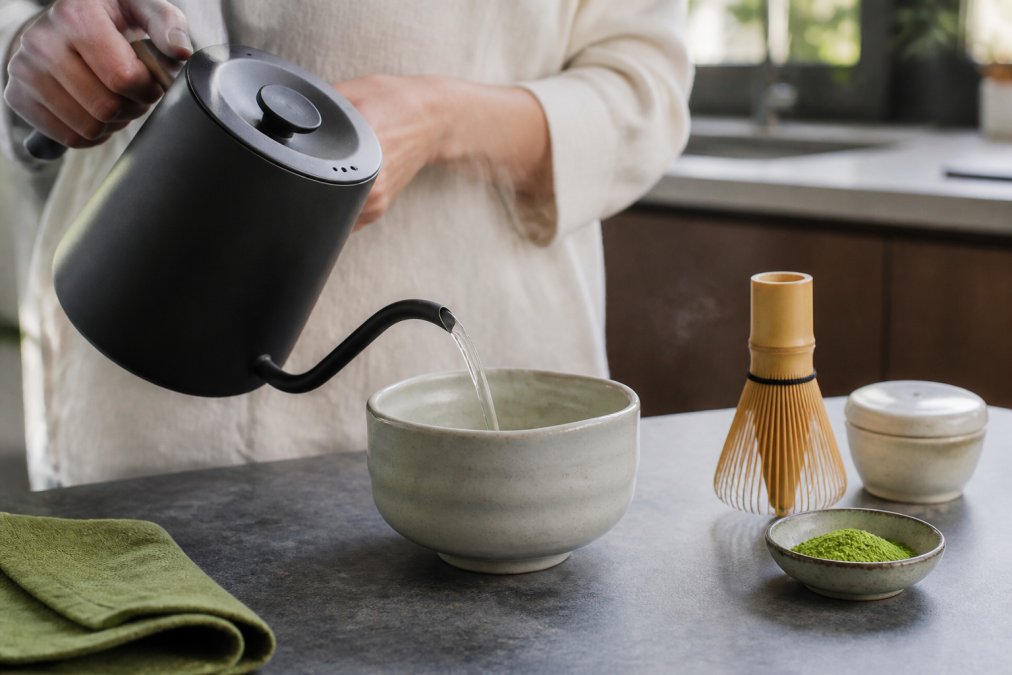 Hands pouring steaming water from a kettle into a small ceramic bowl next to a bamboo whisk and matcha powder