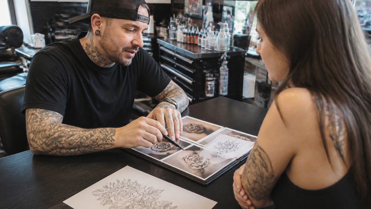Professional tattoo artist and client reviewing reference images and design sketches at consultation table with natural studio lighting