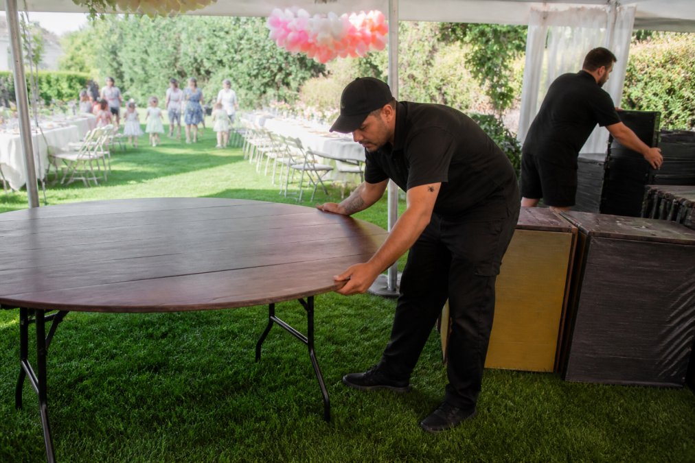 Event rental staff efficiently setting up equipment at an outdoor venue with colorful decorations, while party guests and families arrive in the background on a sunny afternoon