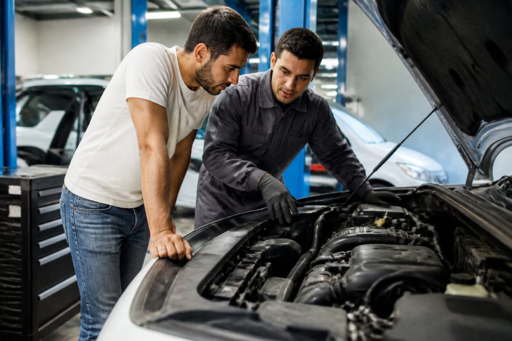 Car owner in casual clothing speaking with professional mechanic wearing work uniform in well-lit modern garage, both examining vehicle engine together with focused attention