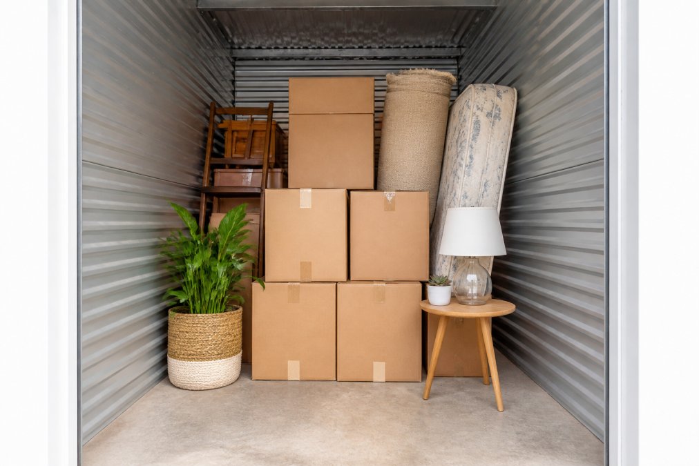 Interior view of a small self-storage unit with neatly stacked cardboard boxes, furniture pieces, and household items arranged to show scale and usable capacity with clear visibility of the space