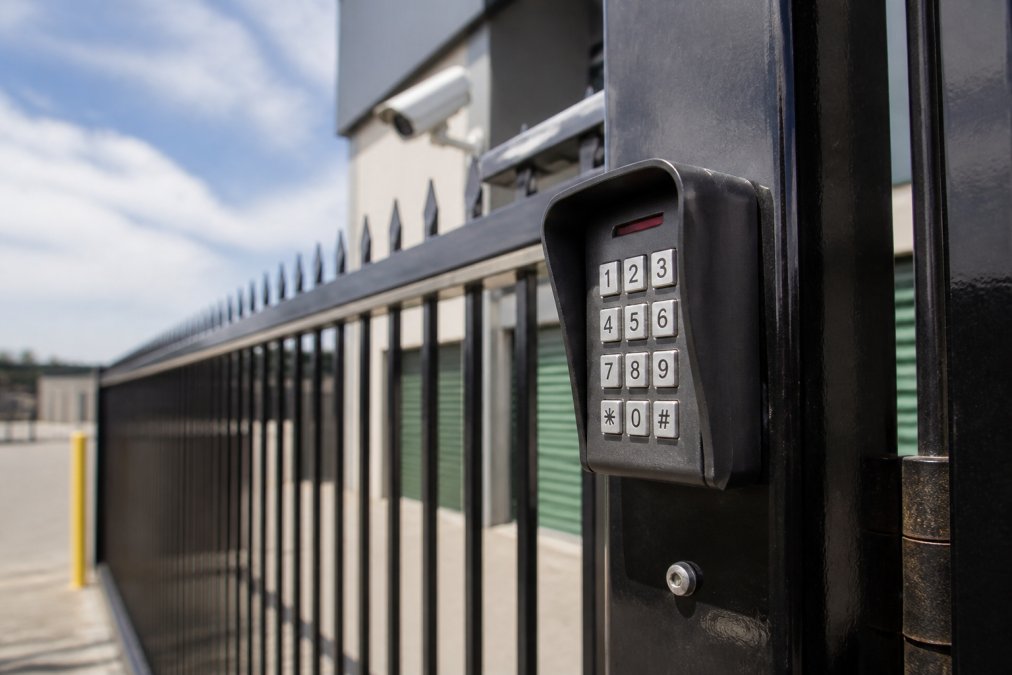 Close-up of a modern keypad PIN entry system mounted on a security gate, with perimeter fencing visible in the background and a CCTV camera mounted on the facility exterior
