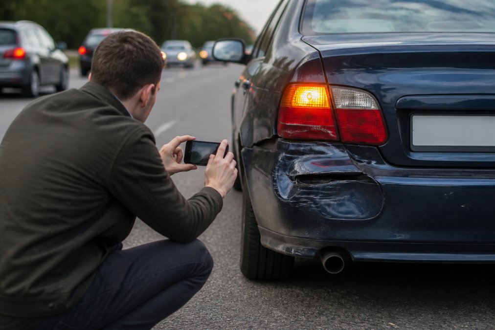Person crouching near a damaged car bumper, holding a smartphone to photograph vehicle damage at the roadside, with hazard lights visible and blurred traffic in the background