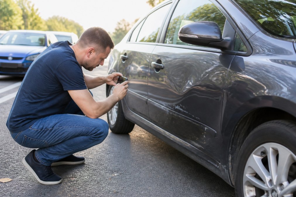 Person crouching beside a damaged car door, photographing the dent and paint damage with a smartphone at the roadside on a clear day