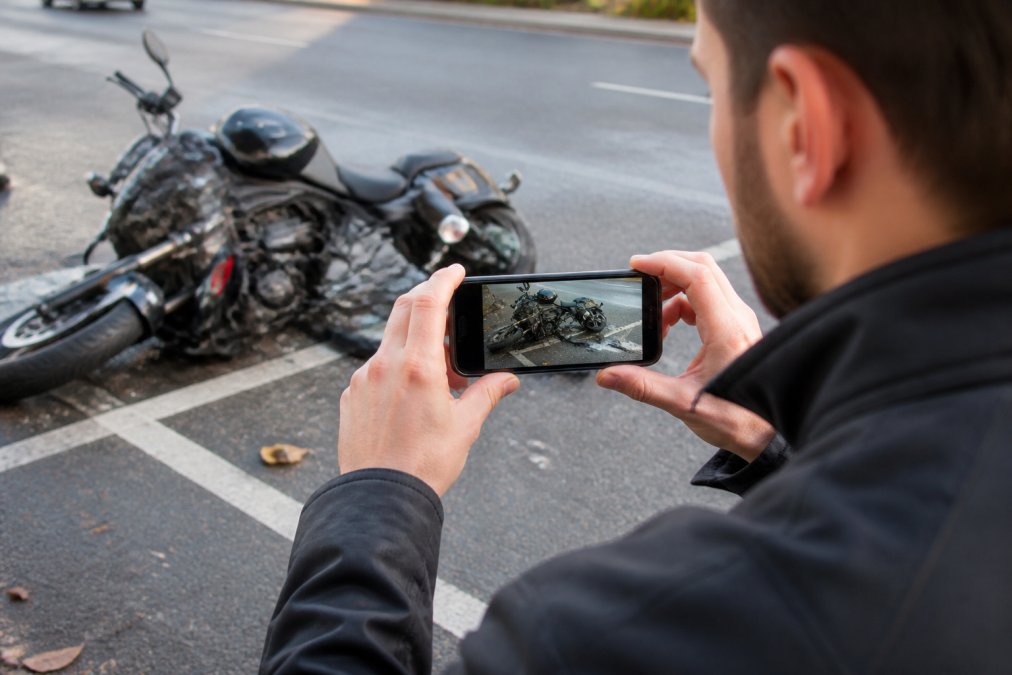 Person using smartphone to photograph motorcycle accident scene with damaged motorcycle and road visible in background, showing close-up detail of damage and surrounding area