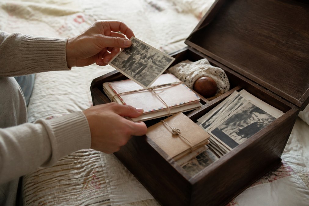 Hands carefully arranging old photographs, handwritten letters, and small mementos into an open wooden memory box with soft natural lighting illuminating the personal items