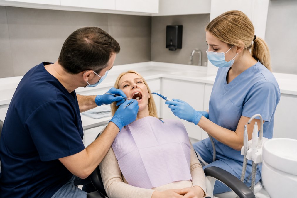 Patient seated in a modern dental chair during routine examination, with dentist and hygienist conducting thorough oral assessment in bright, well-lit clinical surgery with contemporary equipment